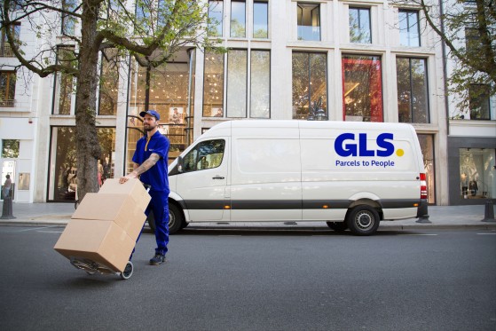 A GLS driver pushes packages on a dolly in front of a GLS van on a city street.